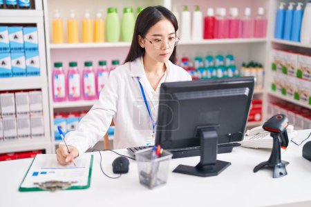 Téléchargez les photos : Young chinese woman pharmacist writing on document using computer at pharmacy - en image libre de droit