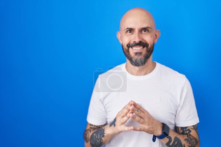 Foto de Hombre hispano con tatuajes de pie sobre fondo azul manos juntas y dedos cruzados sonriendo relajado y alegre. éxito y optimismo - Imagen libre de derechos