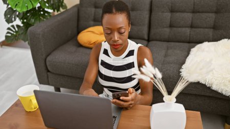 Foto de Mujer afroamericana enfocada usando computadora portátil y teléfono celular en casa, trabajando en su dispositivo en la mañana alimentada con café se sentó atractivamente en el sofá, completamente inmerso en la tecnología, - Imagen libre de derechos