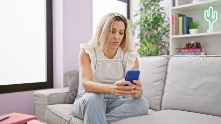 Foto de Mujer rubia joven usando teléfono inteligente sentado en el sofá con la cara seria en casa - Imagen libre de derechos