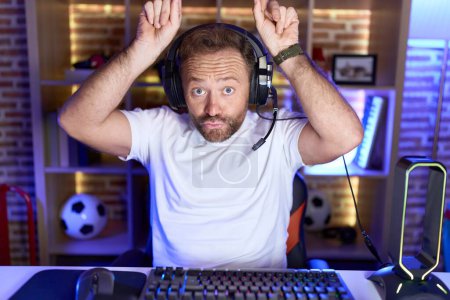 Téléchargez les photos : Homme d'âge moyen avec barbe jouant à des jeux vidéo portant des écouteurs faire geste drôle avec doigt sur la tête comme cornes de taureau - en image libre de droit