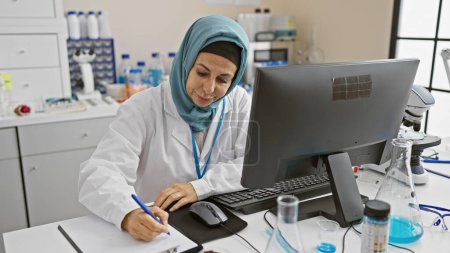 Photo for A professional woman in hijab taking notes in a laboratory setting, surrounded by scientific equipment. - Royalty Free Image