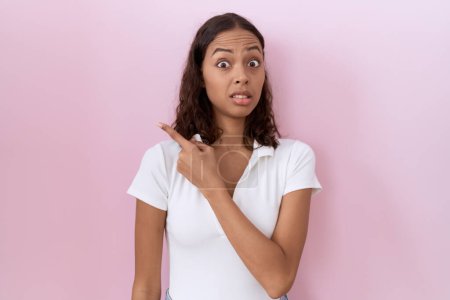 Foto de Mujer hispana joven con camiseta blanca casual señalando a un lado preocupada y nerviosa con el dedo índice, preocupada y sorprendida expresión - Imagen libre de derechos