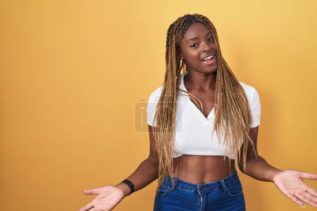 Foto de Mujer afroamericana con el pelo trenzado de pie sobre fondo amarillo sonriendo alegre con los brazos abiertos como bienvenida amistosa, saludos positivos y confiados - Imagen libre de derechos