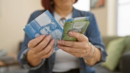 Foto de Mujer hispana que compara pesos chilenos con dólares americanos en el interior, lo que implica consideraciones financieras en el hogar. - Imagen libre de derechos