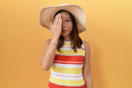 Téléchargez les photos : Femme chinoise d'âge moyen portant un chapeau d'été sur fond jaune couvrant un oeil avec la main, sourire confiant sur le visage et l'émotion surprise. - en image libre de droit
