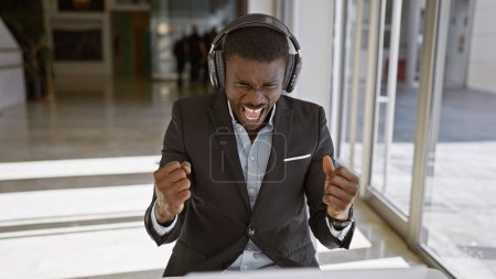 Foto de Emocionado hombre afroamericano en traje de negocios con auriculares celebrando en un entorno de oficina moderno. - Imagen libre de derechos