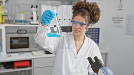 Photo for A young hispanic woman, with curly hair, examines a flask in a lab setting, illustrating science and healthcare. - Royalty Free Image