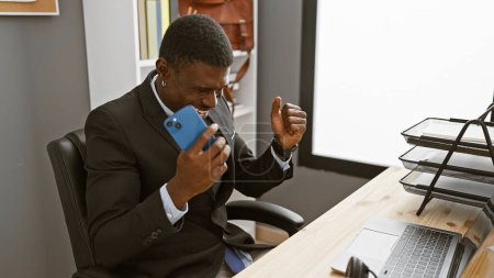 Photo for An african american man in a suit celebrates with a smartphone in an office setting, exuding professionalism and joy. - Royalty Free Image