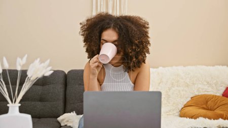 Foto de Una joven con el pelo rizado disfrutando de café en su sala de estar mientras usa un ordenador portátil en el interior. - Imagen libre de derechos