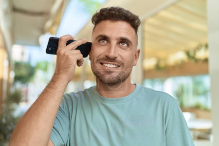 Joven hombre hispano sonriendo confiado escuchando mensaje de audio desde el teléfono inteligente en la terraza de la cafetería