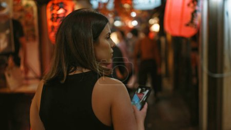 Téléchargez les photos : Belle vue arrière de la femme hispanique alors qu'elle s'éloigne dans l'omoide yokocho de tokyo, smartphone à la main, lors d'une promenade nocturne décontractée. vue sur la rue capturer ses cheveux bruns et la vie vibrante de la ville. - en image libre de droit