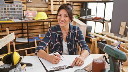 Téléchargez les photos : Une femme souriante dessine des plans dans un atelier de menuiserie bien équipé entouré d'outils et de bois. - en image libre de droit