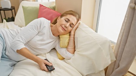 Téléchargez les photos : Femme caucasienne mature se détendre dans la chambre à coucher avec télécommande - en image libre de droit