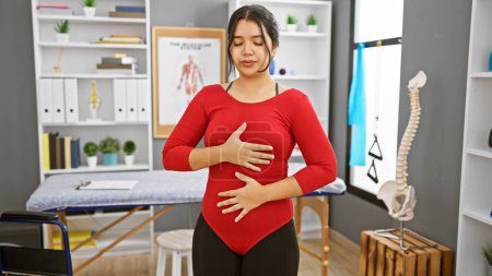 Hispanic woman practicing breathing exercises in a clinic interior with a visible spine model.
