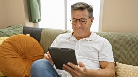 Foto de Un hombre de mediana edad se relaja en casa, interactuando con una tableta en su acogedora sala de estar. - Imagen libre de derechos