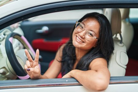 Téléchargez les photos : Young chinese woman driving car doing victory gesture with fingers at street - en image libre de droit