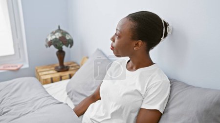 Attraktive afrikanisch-amerikanische Frau, die in ihrem gemütlichen Bett erwacht, mit ernstem Gesichtsausdruck in ihrem Schlafzimmer sitzt und die Morgendämmerung betrachtet.