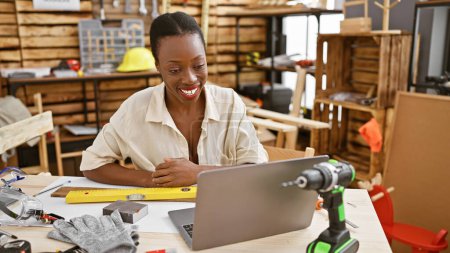Téléchargez les photos : Belle femme charpentier afro-américaine souriante alors qu'elle travaille sur son entreprise de menuiserie avec ordinateur portable à l'atelier de menuiserie - en image libre de droit