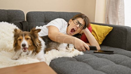Téléchargez les photos : Une jeune femme hispanique bâillant et un chien se relaxant sur un canapé dans un salon confortable. - en image libre de droit