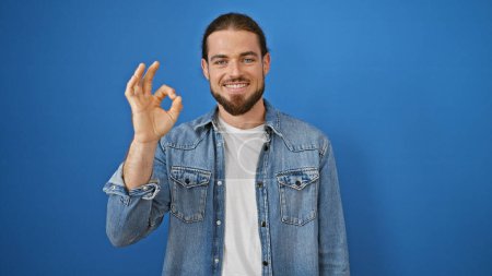 Téléchargez les photos : Jeune homme hispanique souriant confiant faire un geste correct sur fond bleu isolé - en image libre de droit