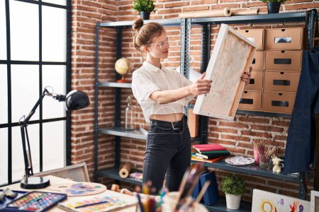 Foto de Mujer rubia examina obras de arte en un interior de estudio creativo, rodeado de materiales de pintura y unidades de estanterías. - Imagen libre de derechos