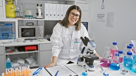 Photo for Smiling hispanic woman scientist in lab coat working in laboratory with microscope and flasks - Royalty Free Image