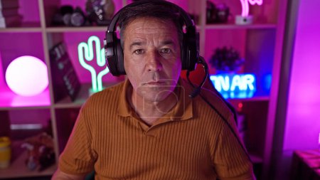 Photo for Middle-aged man with headphones in a colorful neon-lit gaming room at night. - Royalty Free Image