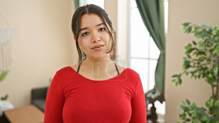 Téléchargez les photos : Une jeune femme hispanique en chemise rouge pose en toute confiance dans un salon moderne, transmettant une élégance décontractée. - en image libre de droit