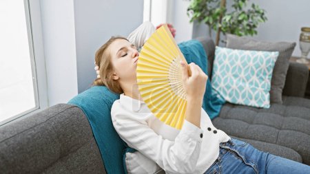 Foto de Una joven se sienta en una moderna sala de estar, enfriándose con un ventilador amarillo mientras se sienta cómodamente en un sofá azul. - Imagen libre de derechos