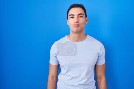 Photo for Young hispanic man standing over blue background looking at the camera blowing a kiss on air being lovely and sexy. love expression. - Royalty Free Image