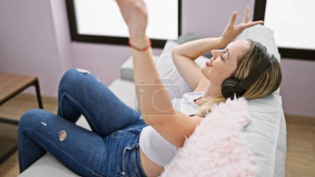 Téléchargez les photos : Jeune femme blonde écoutant de la musique assise sur un canapé dansant à la maison - en image libre de droit