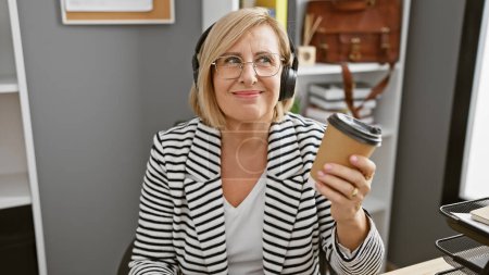 Téléchargez les photos : Femme souriante portant des écouteurs dans un blazer rayé tient café pendant le travail de bureau. - en image libre de droit