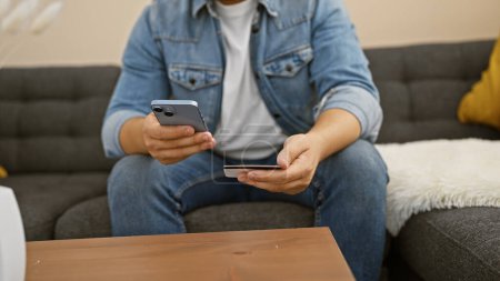 Foto de Un hombre que usa una tarjeta de crédito y un teléfono inteligente para ir de compras en línea mientras está sentado en un sofá en su sala de estar. - Imagen libre de derechos