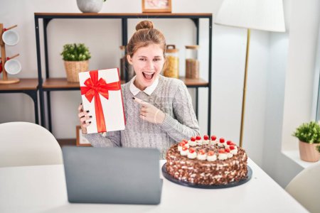Téléchargez les photos : Jeune femme blonde excitée célèbre joyeusement son anniversaire à la maison, pointant joyeusement son cadeau tout en rayonnant avec un sourire, mettant en valeur le pur bonheur sur un appel vidéo d'ordinateur portable. - en image libre de droit