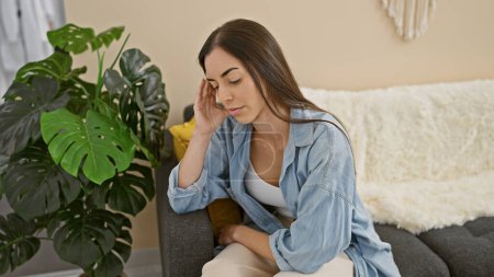 Photo for Portrait of a young, beautiful hispanic woman expressing worry, doubt and serious thoughts while sitting indoors on a living room sofa at home, visibly tired, a pensive look on her attractive face - Royalty Free Image