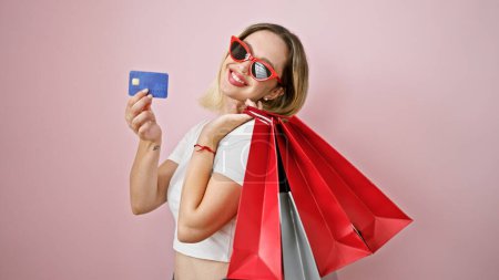 Foto de Joven mujer rubia cliente sosteniendo bolsas de compras y tarjeta de crédito sobre fondo rosa aislado - Imagen libre de derechos