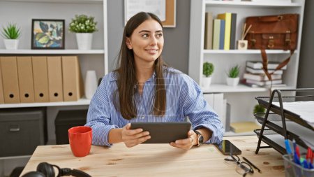 Foto de Una mujer confiada disfruta de un momento en el trabajo, sosteniendo una tableta en una oficina moderna. - Imagen libre de derechos