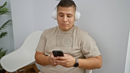 Téléchargez les photos : Jeune homme latino utilisant smartphone et écouteurs assis sur la chaise dans la salle d'attente - en image libre de droit