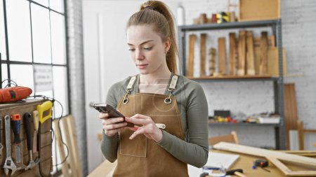 Foto de Una joven enfocada en un taller, usando un delantal, usando un teléfono inteligente en medio de herramientas de carpintería. - Imagen libre de derechos