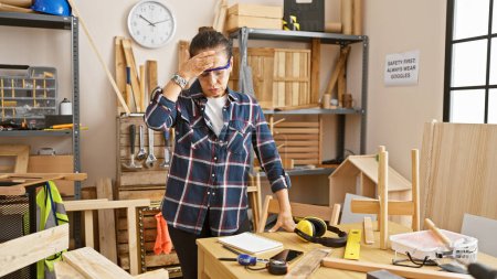 Photo for Stressed woman in workshop with safety goggles pauses amid woodwork tools and unfinished projects. - Royalty Free Image
