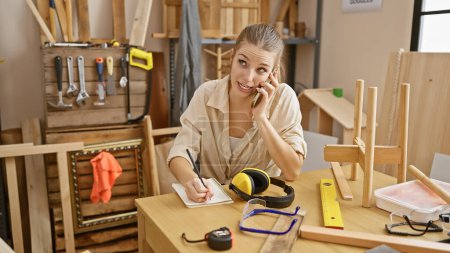 Foto de Una joven mujer multitarea en un taller de carpintería, hablando por teléfono mientras escribe notas en medio de varias herramientas. - Imagen libre de derechos