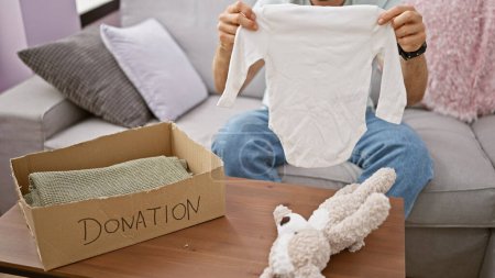 Young hispanic man sitting on sofa packing clothes and teddy bear on cardboard box to donate at home