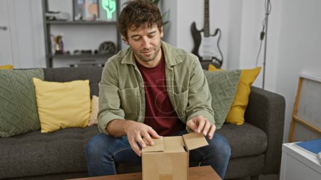 Foto de Un joven casual con barba desempacando una caja de cartón en un acogedor apartamento sala de estar. - Imagen libre de derechos