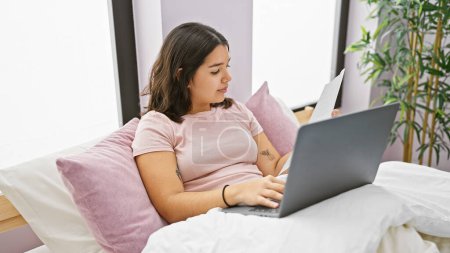 Young hispanic woman working on laptop in bedroom with a relaxed posture and comfortable interior
