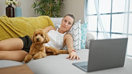Téléchargez les photos : Jeune femme caucasienne avec chien regarder film sur ordinateur portable couché sur canapé souriant à la maison - en image libre de droit
