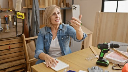 Foto de Una mujer mayor tomando una selfie en un taller de carpintería con herramientas y anteojos sobre la mesa. - Imagen libre de derechos