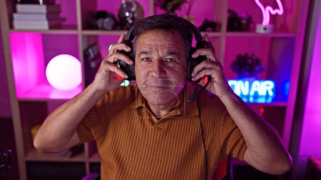 Photo for Mature man enjoys gaming at night in a neon-lit room, donning headphones with a playful smile. - Royalty Free Image