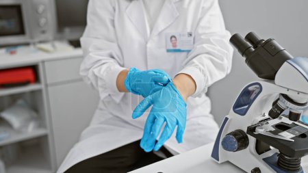 Foto de Una mujer científica con bata blanca se pone guantes azules en un moderno entorno de laboratorio. - Imagen libre de derechos
