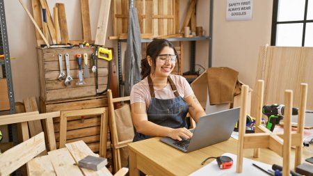 Photo for A smiling hispanic woman works on a laptop in a woodwork workshop surrounded by tools and lumber. - Royalty Free Image
